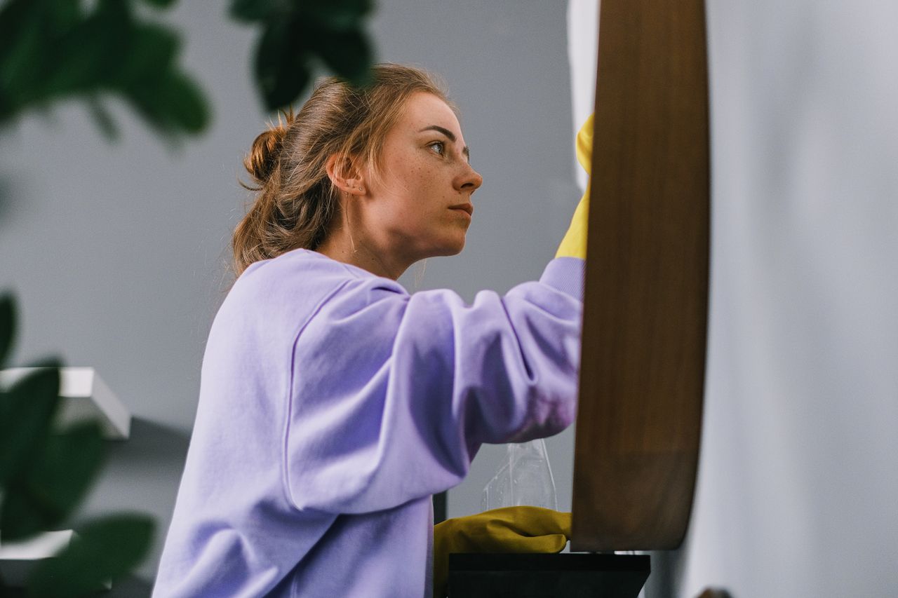 Vue de côté d’une jeune femme concentrée, portant des gants en latex, debout et nettoyant un miroir dans une pièce lumineuse en journée